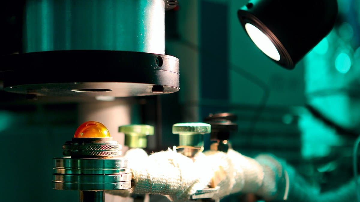 Close-up of the sample stage of a diffuse-reflectance infrared spectrometer, an amber sample dome and knurled adjusters lit under a lamp in a laboratory.