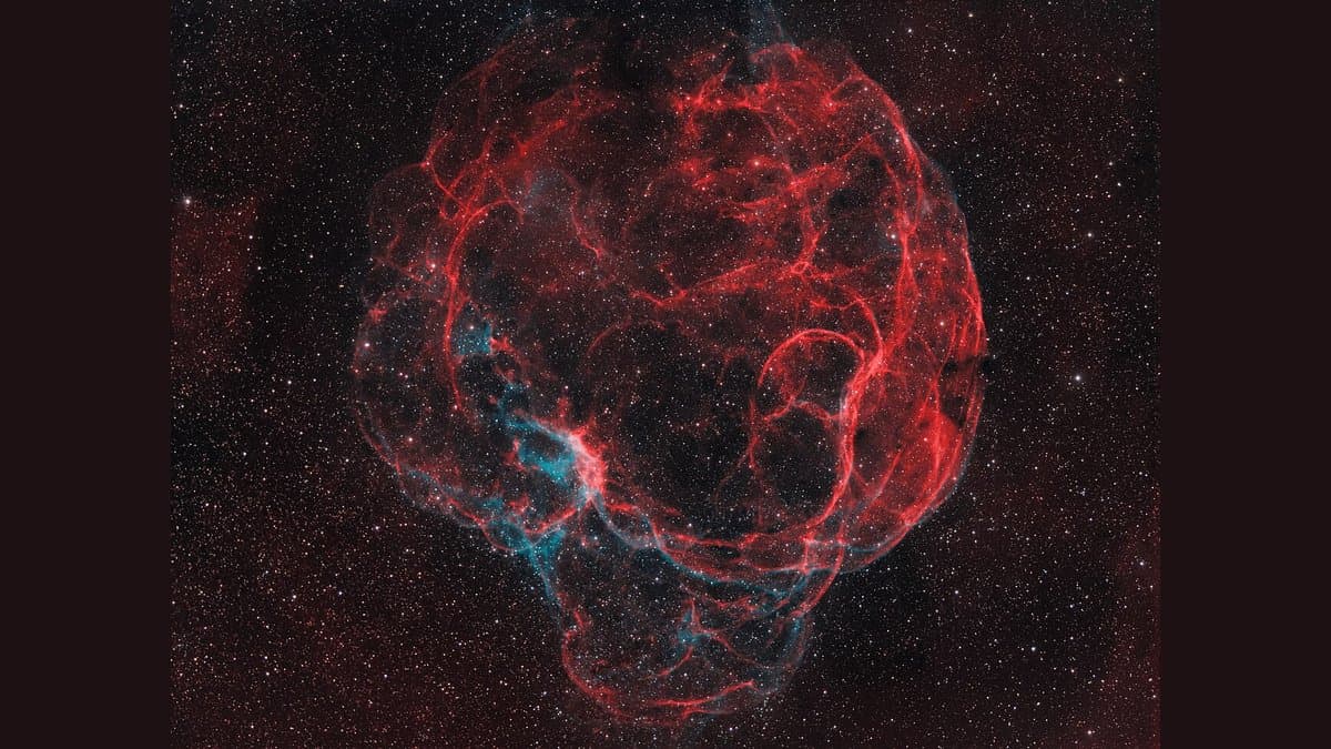 A large, faint supernova remnant filling the frame: a rough sphere of tangled red hydrogen filaments laced with blue-green oxygen emission, set against a dense field of stars.
