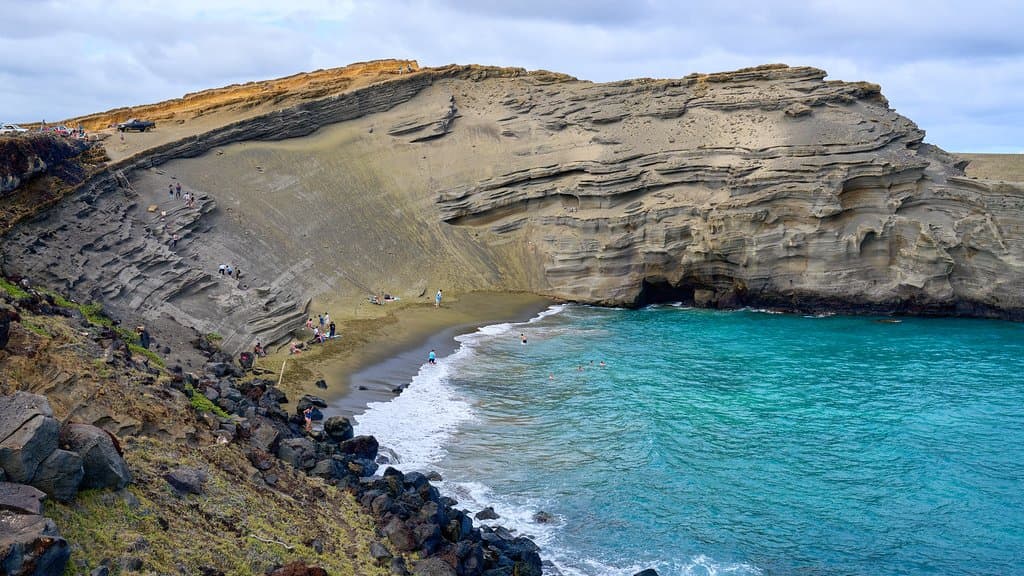 A bay of olive-green sand enclosed by dark volcanic cliffs, with surf breaking on the shore at Papakolea Green Sand Beach, Hawaii.