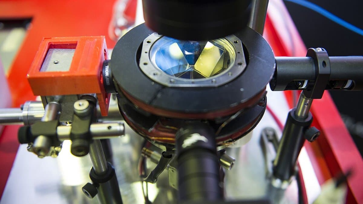 Close-up of an atom interferometer: a vacuum chamber ringed with coils and windows, surrounded by laser optics on a laboratory mount.