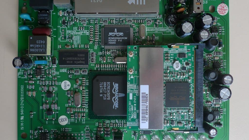 The bare green circuit board of a consumer wireless router, lifted out of its case, showing two large chips, memory, rows of capacitors and a metal four-port Ethernet block.