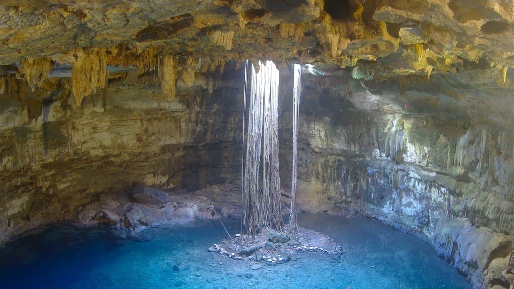 A cavern pool of clear blue groundwater below a limestone roof, lit by daylight falling through an opening above, at Cenote Dzitnup in Yucatan, Mexico.