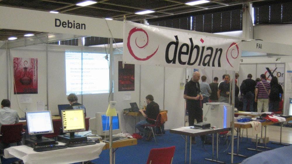 A Debian Project exhibition stand under a large white banner carrying the debian wordmark and swirl logo, with volunteers and desktop computers on tables.