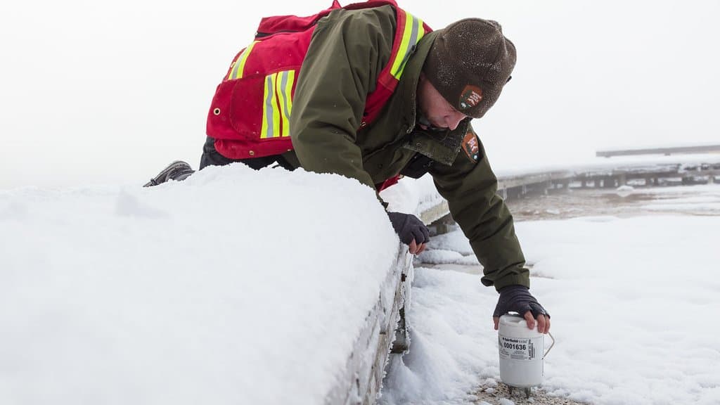 A geologist in winter gear kneels on a snow-covered boardwalk and lowers a small cylindrical nodal seismometer into frozen ground.