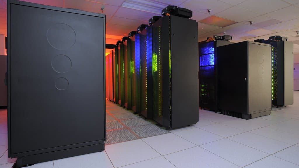 Rows of tall black computer cabinets lit from within by blue and green indicator lights, standing on a white tiled floor in a machine room.