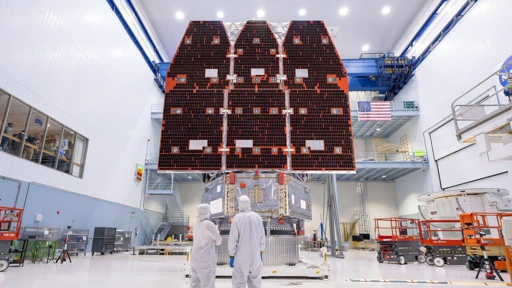 The Nancy Grace Roman Space Telescope in a clean room at NASA's Goddard Space Flight Center, its six-panel Solar Array Sun Shield mounted above the spacecraft bus, with two technicians in white coveralls standing beneath it for scale.
