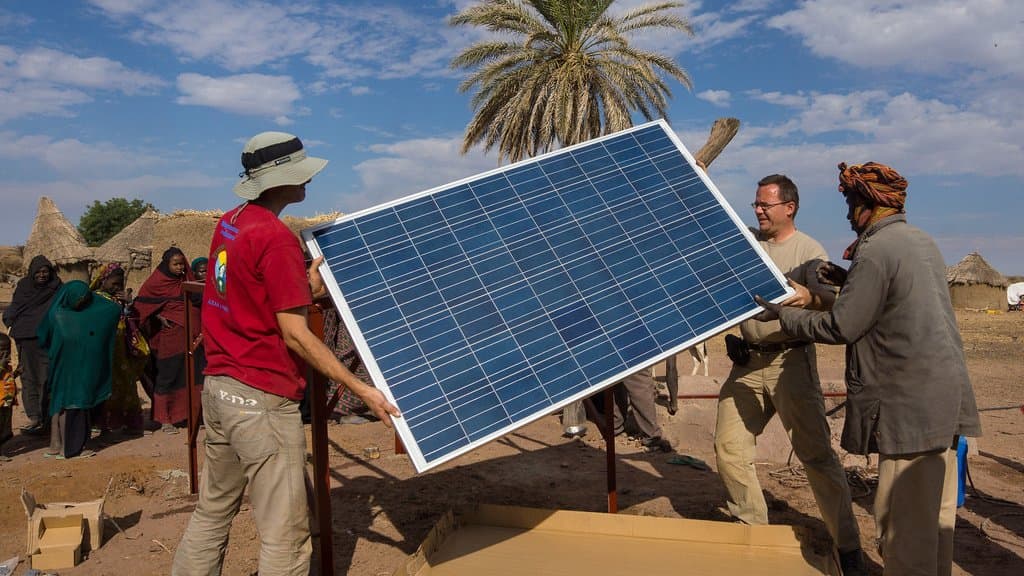 Several people lifting a large blue photovoltaic panel onto a frame in a village of thatched huts under a palm tree, in a dry Sahelian landscape.