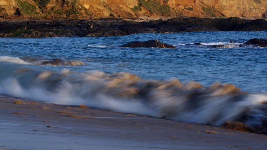A wave breaking and running up a sandy beach in the foreground, with dark rocks and a cliff face lit by low sun behind.