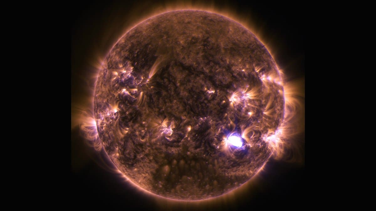 The full disk of the Sun imaged in extreme ultraviolet light, showing a mottled orange surface laced with magnetic loops and one intensely bright white flare near the lower right edge.