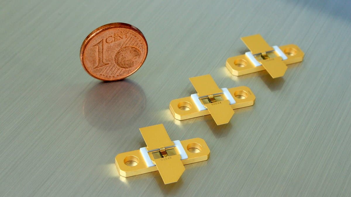 Three small gold-plated packaged gallium nitride power transistors lying on a brushed metal surface beside a one-cent coin for scale.