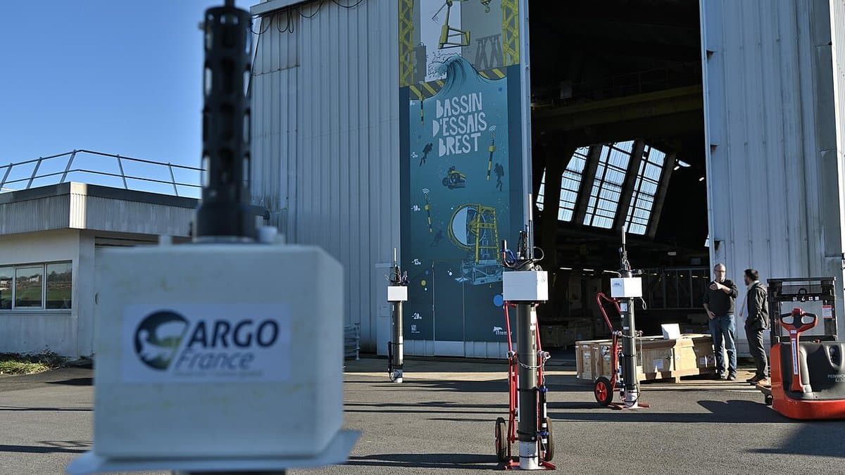 A row of upright cylindrical Argo profiling floats on wheeled trolleys outside a large test-basin hangar, with an Argo France instrument label in the foreground.