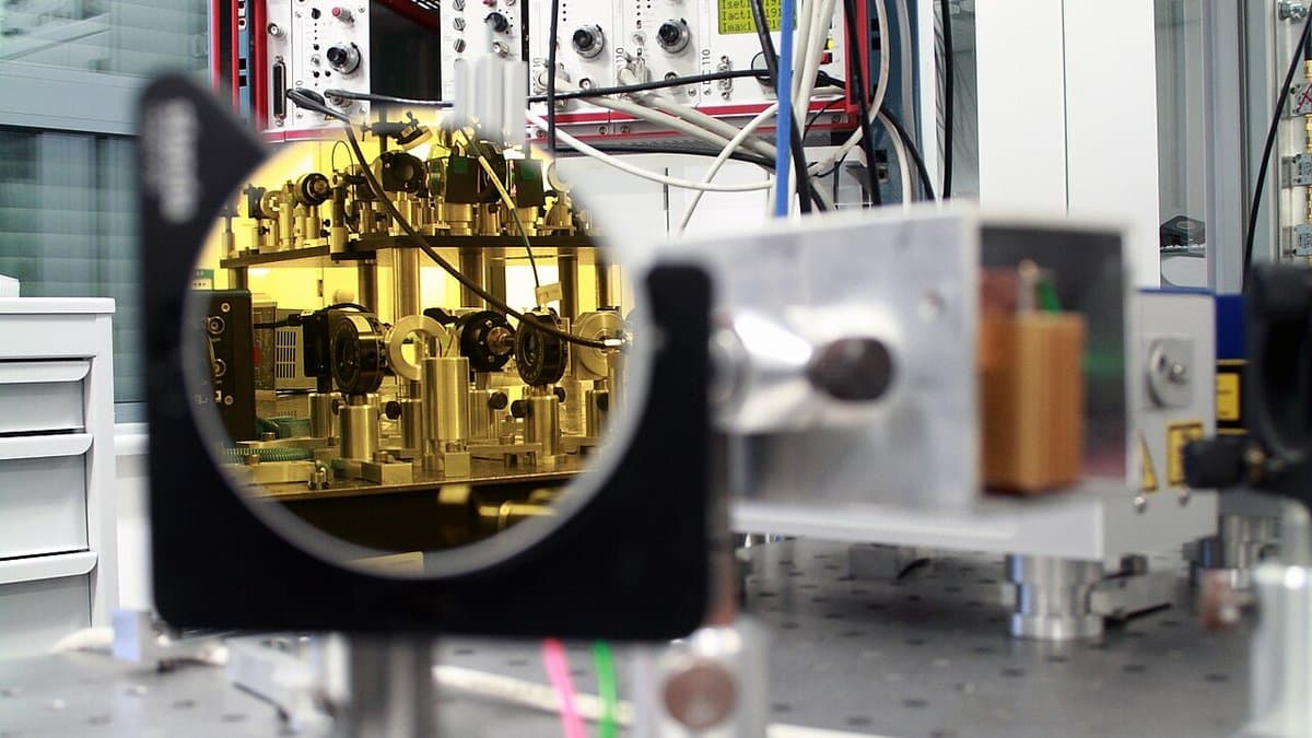 A quantum optics bench: a mirror mounted on an optical table reflects a gold-plated vacuum chamber assembly, with electronics racks behind it.