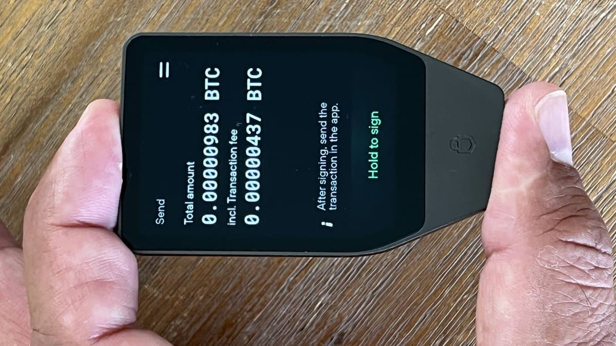A pair of hands holds a black Trezor hardware wallet whose screen shows a bitcoin transaction awaiting on-device confirmation.