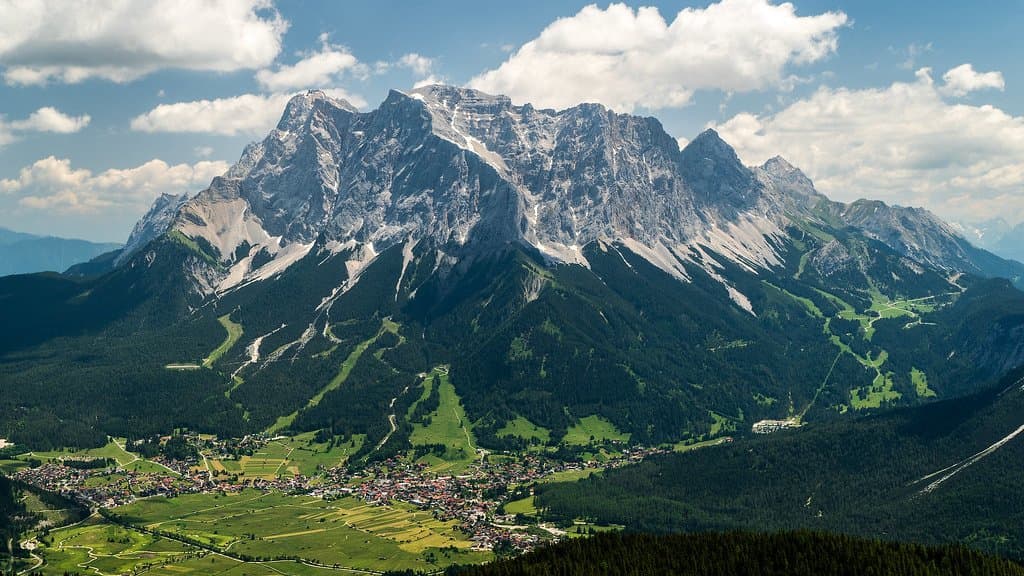 The Zugspitze massif rising above a green alpine valley and village, its grey limestone faces streaked with old snow under a partly cloudy sky.