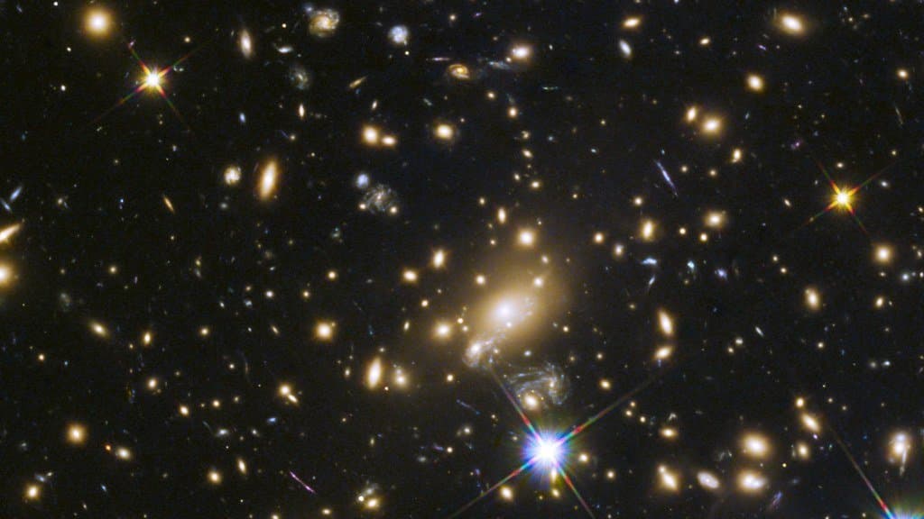 Hubble image of galaxy cluster MACS J1149.6+2223: a dense field of yellow elliptical galaxies and faint blue arcs of stretched background galaxies against black sky.