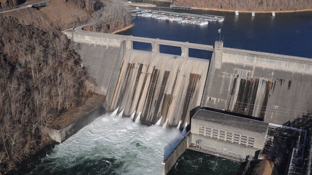 Aerial view of a concrete hydroelectric dam releasing water through spillway gates, with a powerhouse at its base and a reservoir stretching away behind the dam wall.