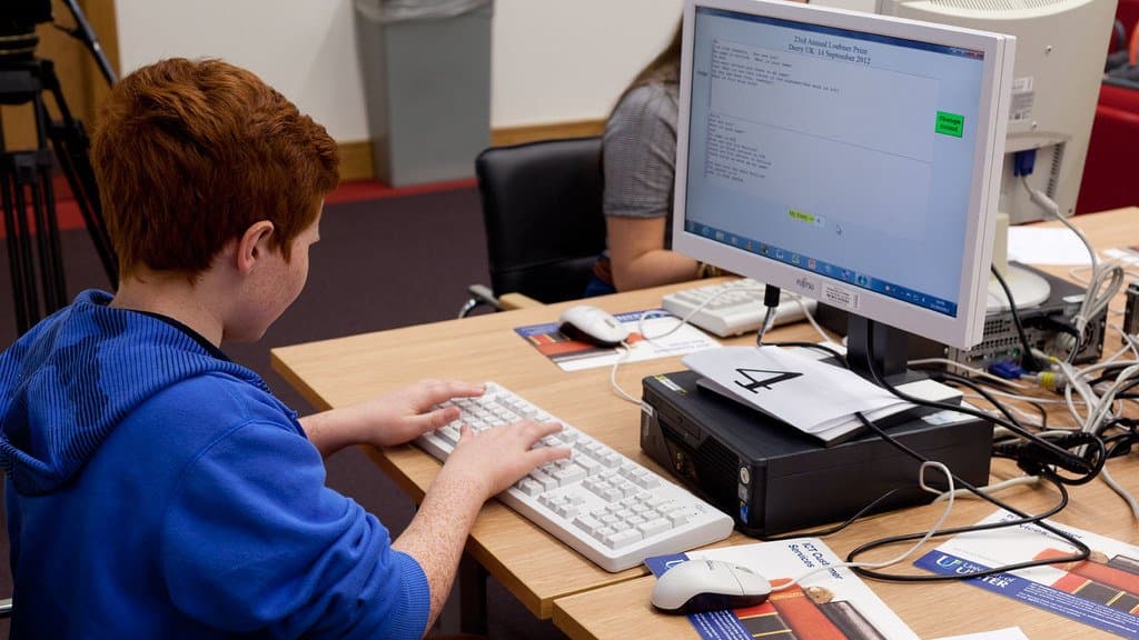 A school-age contestant in a hooded top types at a keyboard while reading a chat transcript on a monitor headed 23rd Annual Loebner Prize, Derry UK, 14 September 2013.
