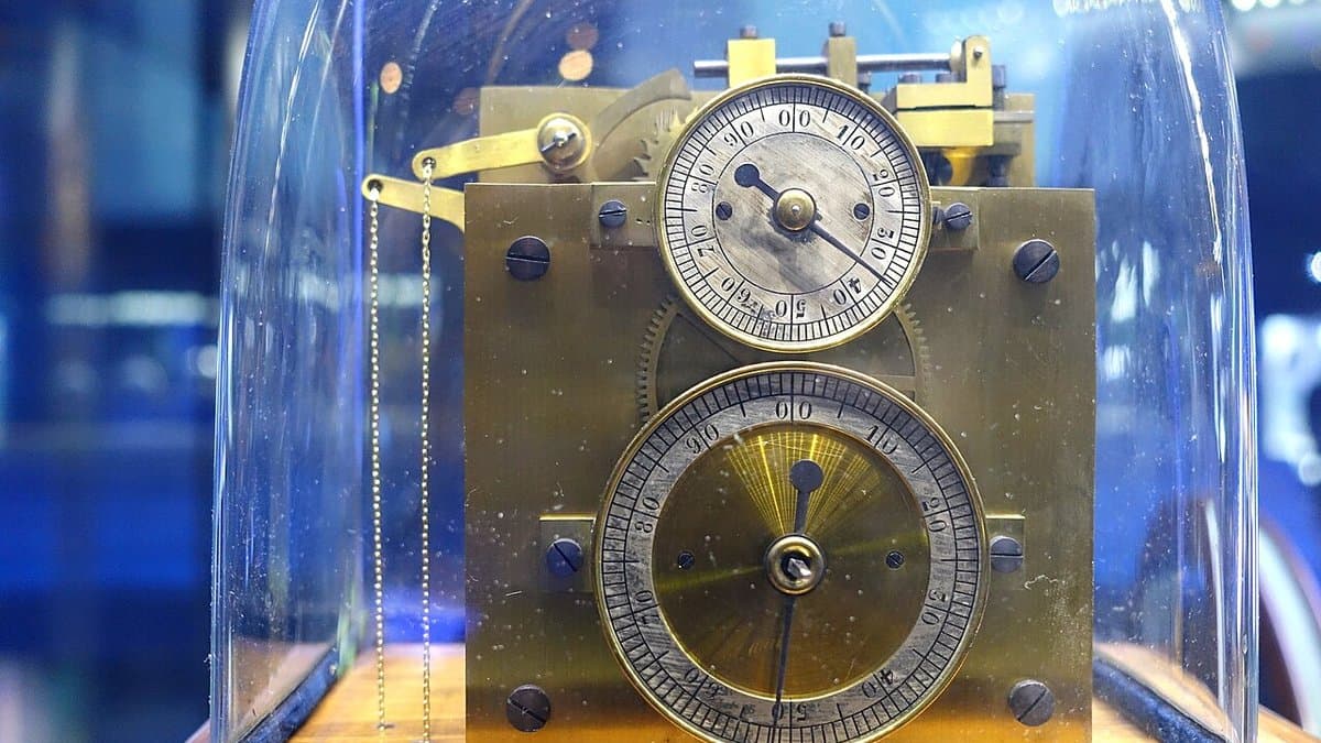 A brass Hipp chronoscope of about 1890 under a glass dome, its two graduated dials marked in thousandths of a second, displayed in a museum case.