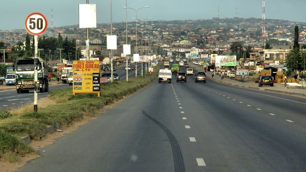 A multi-lane road on the edge of Accra carrying trucks, minibuses and cars, with roadside kiosks and advertising boards on the left and densely packed low-rise houses covering the hillside behind.
