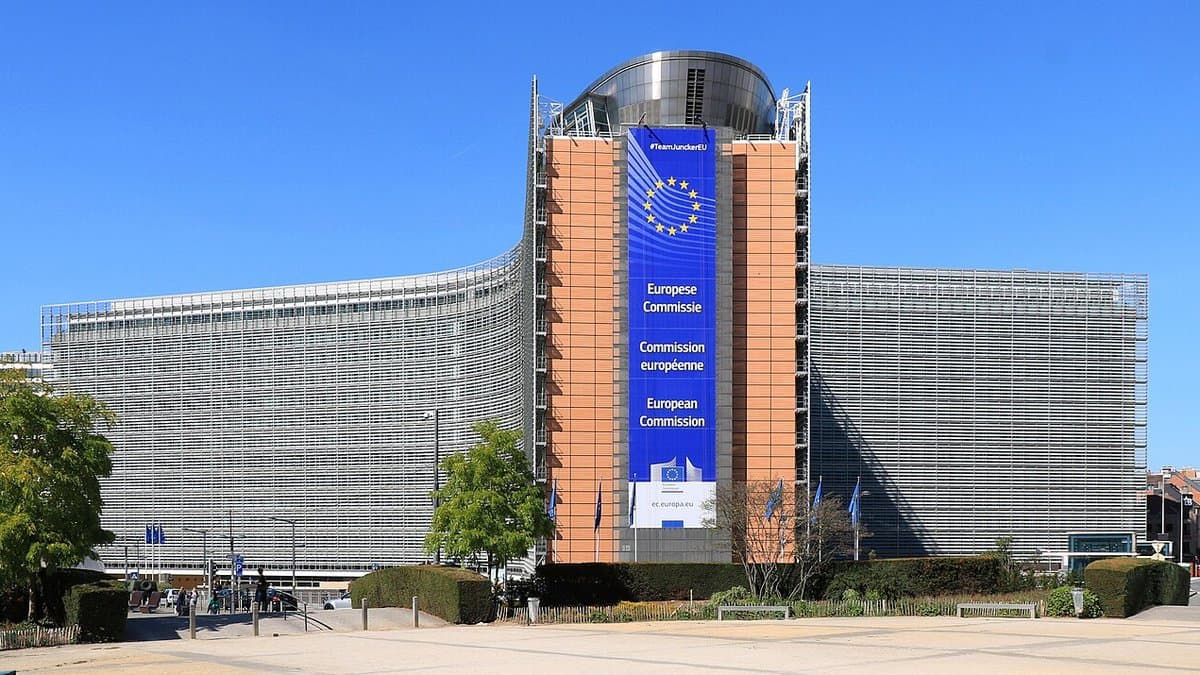 The Berlaymont building in Brussels, a curved glass-and-steel office block, with a large blue banner reading European Commission in Dutch, French and English hung on its central tower beneath a circle of gold EU stars.