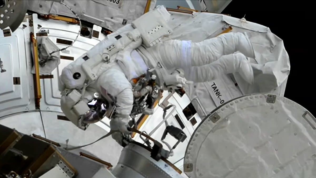 An astronaut in a white spacesuit moves along the exterior of the International Space Station, holding a handrail beside a white module and equipment covers, with black space behind.