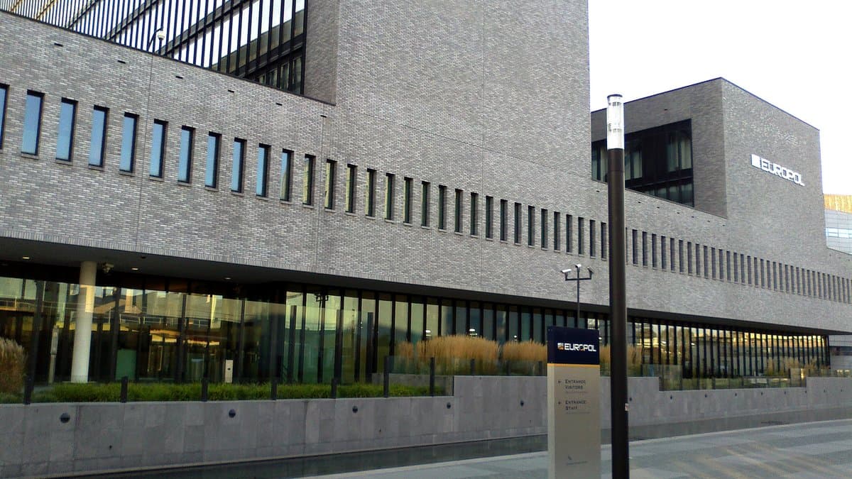 The Europol headquarters in The Hague: a modern brick-and-glass office block with the Europol name mounted on its facade and a signpost at the entrance.