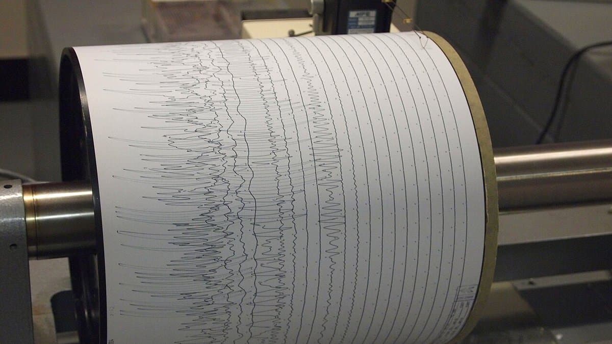 Close-up of a rotating drum seismograph with paper wrapped around it, covered in rows of inked seismic traces including dense bursts of rapid oscillation.