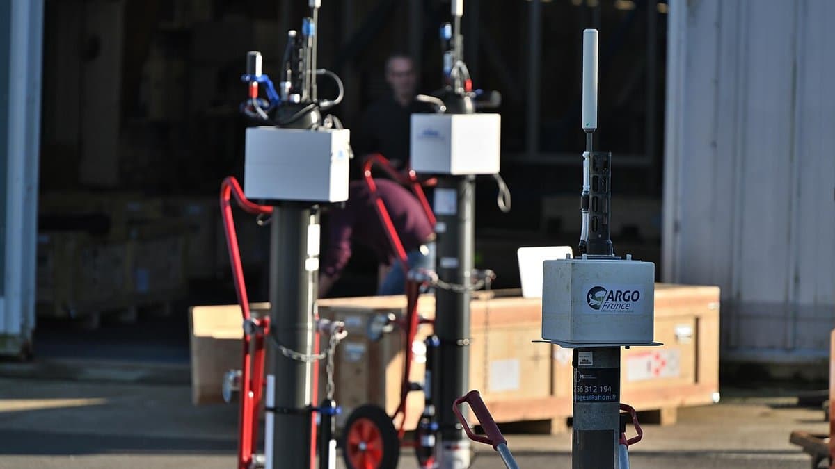 Three tall grey Argo profiling floats with white instrument housings and antennas stand on a quayside in front of an open workshop door, one carrying an ARGO France label.