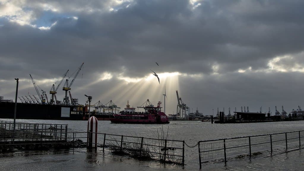 Floodwater covers a fenced riverside promenade in the Port of Hamburg during a storm surge, with container cranes, a ferry and sunbeams breaking through heavy cloud behind.