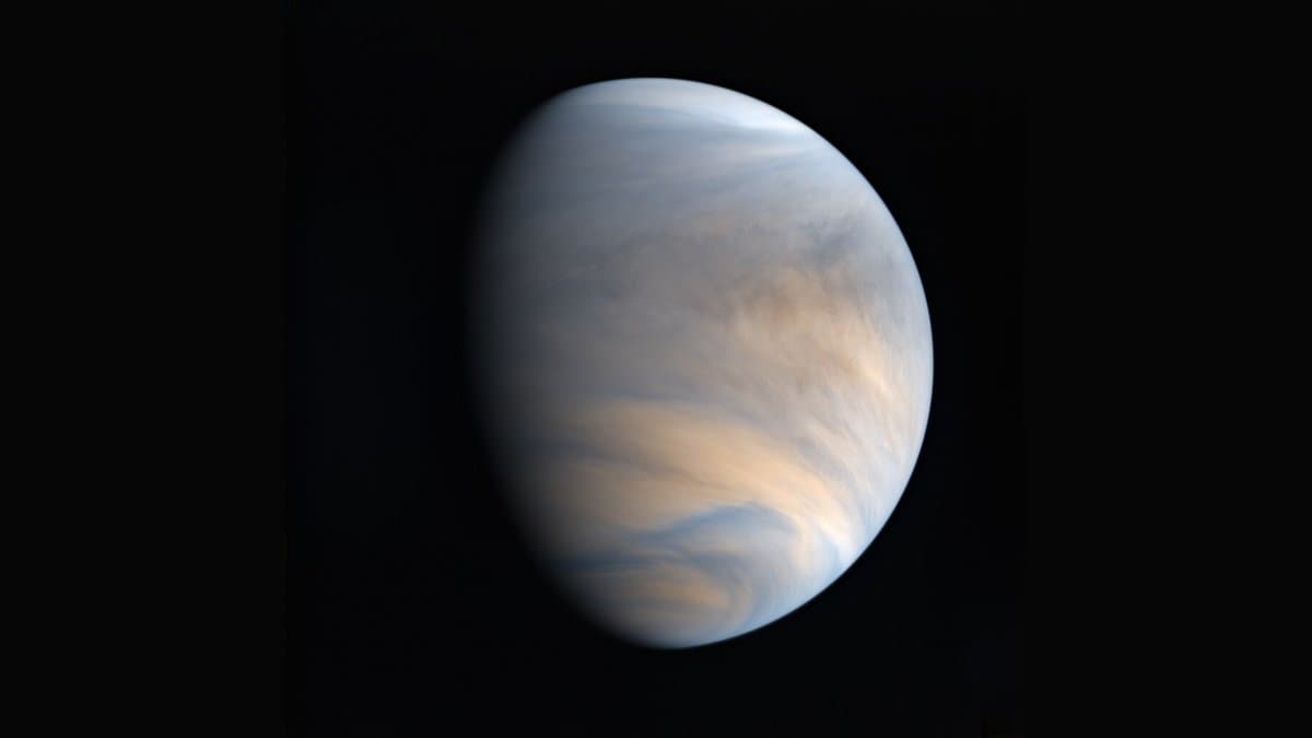 The full disk of Venus, its thick cloud deck banded in cream and pale blue, against black space, imaged by the Akatsuki orbiter.