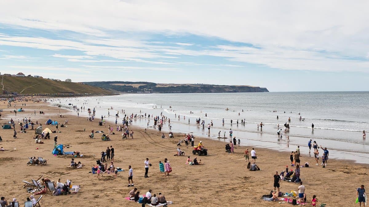 Crowds of people on the wide sandy beach at Whitby, North Yorkshire, on a warm summer day, with the sea on the right and cliffs in the distance.