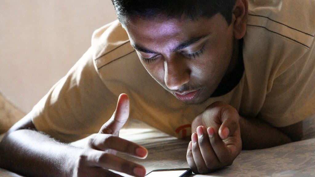 A teenage boy lies on a bed reading a smartphone at close range, his face lit by the screen.
