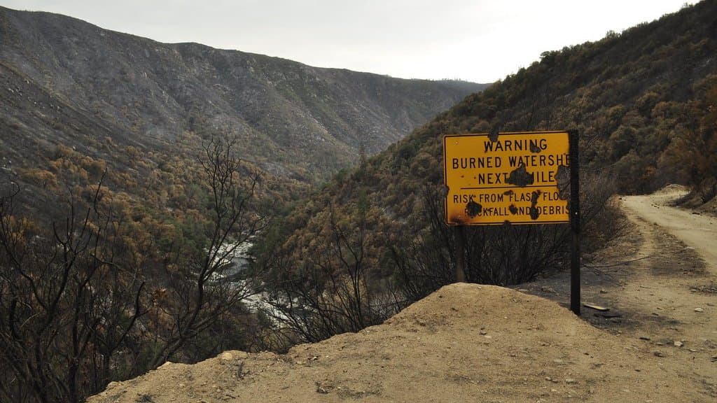 A yellow roadside sign reading Warning: Burned Watershed Next Miles, Risk from Flash Flood, Rockfall and Debris, standing on a dirt track above a steep burned canyon with a river running along its floor.