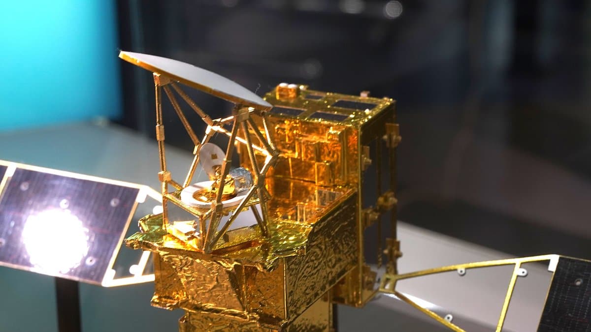 A gold-foil scale model of the GOSAT-GW satellite in a museum display case, with a large white offset dish antenna above the spacecraft body and solar arrays extending to either side.