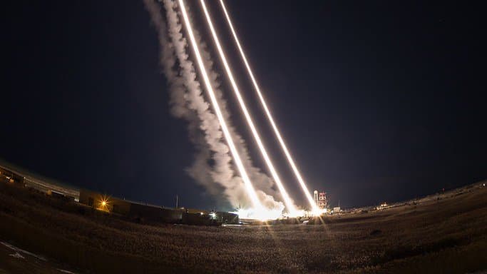 Long-exposure night photograph of three bright rocket trails arcing up from a floodlit launch complex at NASA Wallops Flight Facility, with lit smoke plumes against a black sky.