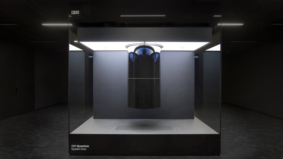 An IBM Quantum System One: a cylindrical cryostat suspended inside a sealed glass cube in a darkened room, with the IBM Quantum System One nameplate on the plinth.