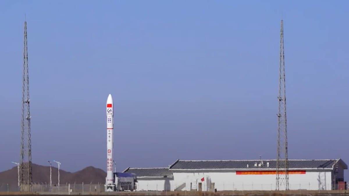 A slender white rocket with red markings stands upright on a launch pad in flat, dry terrain, flanked by two tall lattice lightning towers, with a long low building alongside.