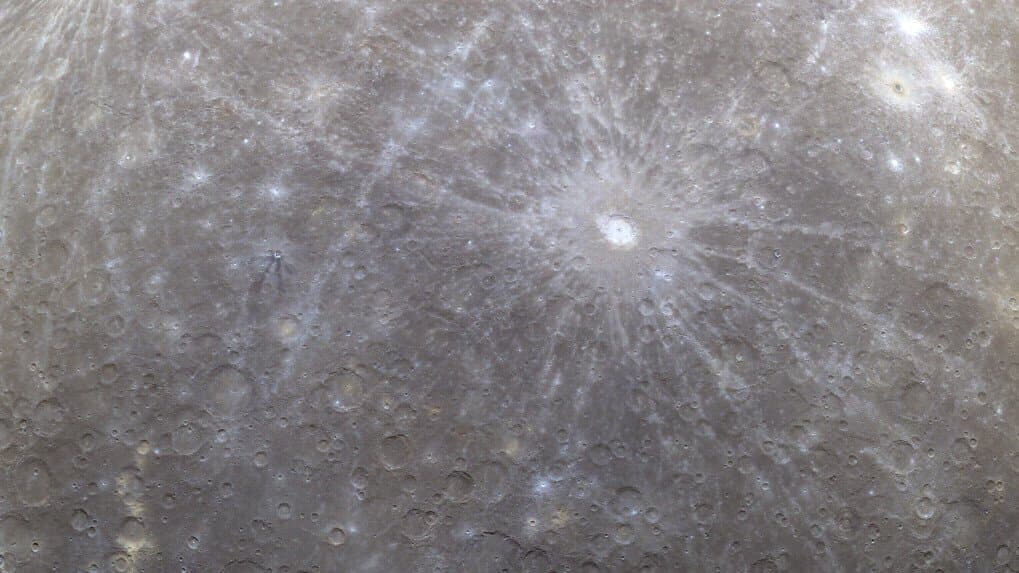 Mercury's gray, heavily cratered surface fills the frame in an orbital view, with a bright rayed impact crater and long, pale rays stretching across older cratered plains.