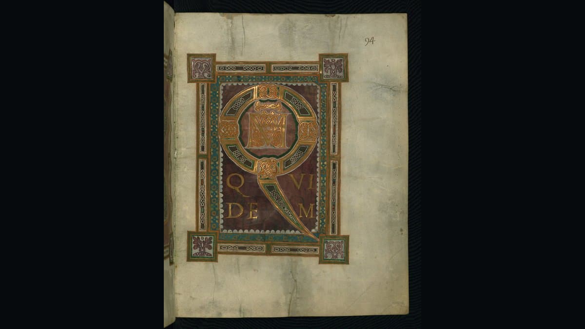 An illuminated medieval manuscript page on parchment: a large gilded initial framed by a jewelled interlace border on a cream vellum leaf.