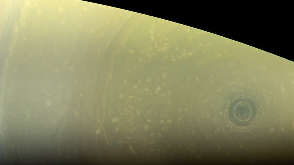 Close-up of Saturn's pale yellow southern hemisphere curving against black space, with a small dark circular storm eye near the pole at right.