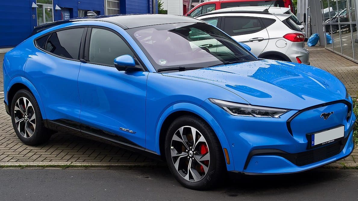 A blue Ford Mustang Mach-E all-electric SUV parked at a kerb, photographed from the front three-quarter angle.