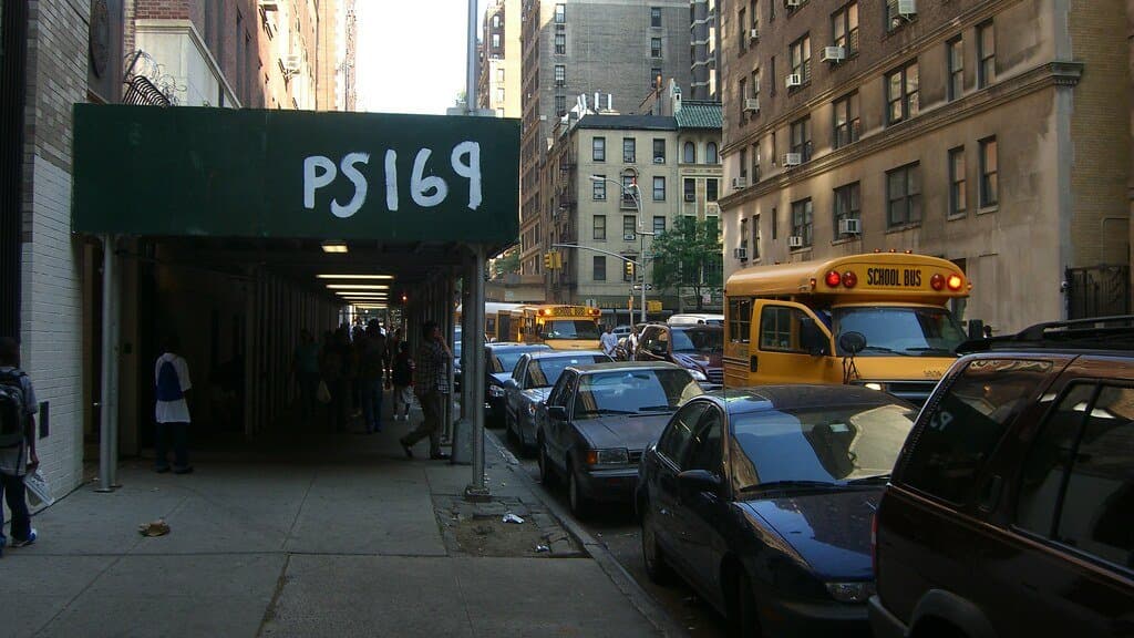 Street entrance of a New York City public school marked P.S. 169, with students under the awning and yellow school buses parked along the block