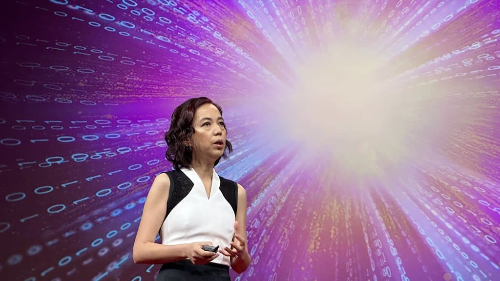 Computer scientist Fei-Fei Li speaks on a stage in front of a large projected display of cascading binary digits.