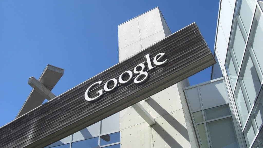A large white Google wordmark on a dark angled beam above the glass entrance of an office building, photographed from below against a clear blue sky.