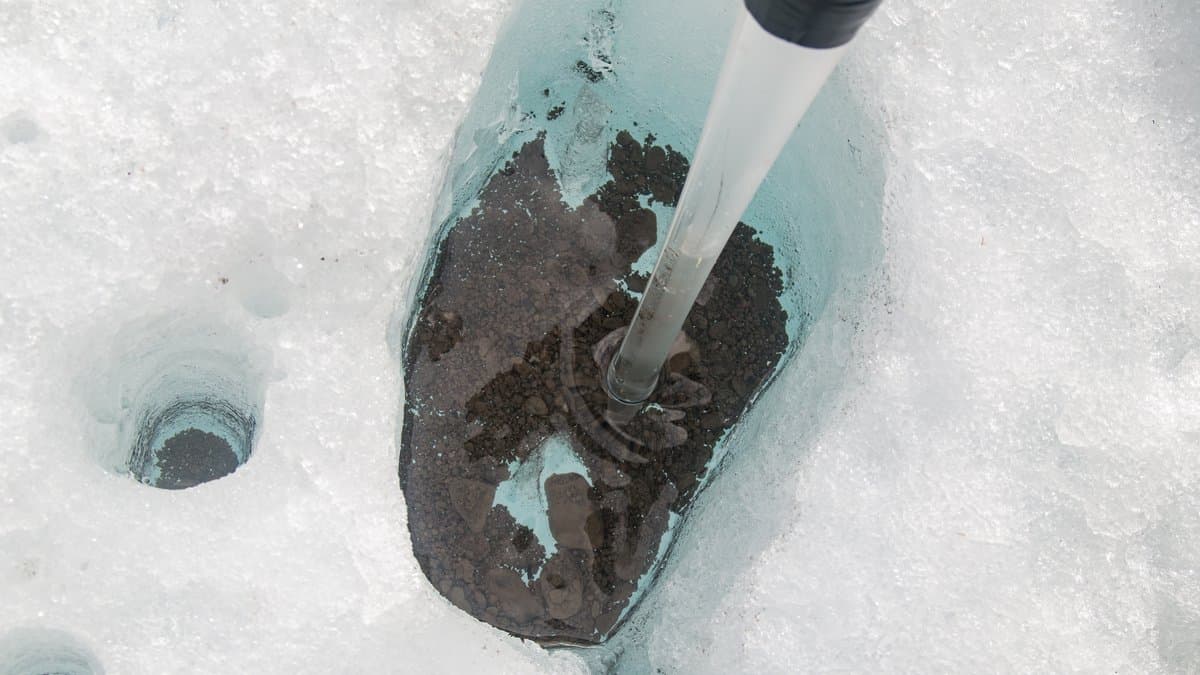Close-up of a glacier surface: an oval meltwater hole in white ice filled with dark grey-brown cryoconite sediment, a sampling pipette lowered into it, with two smaller water-filled holes nearby.