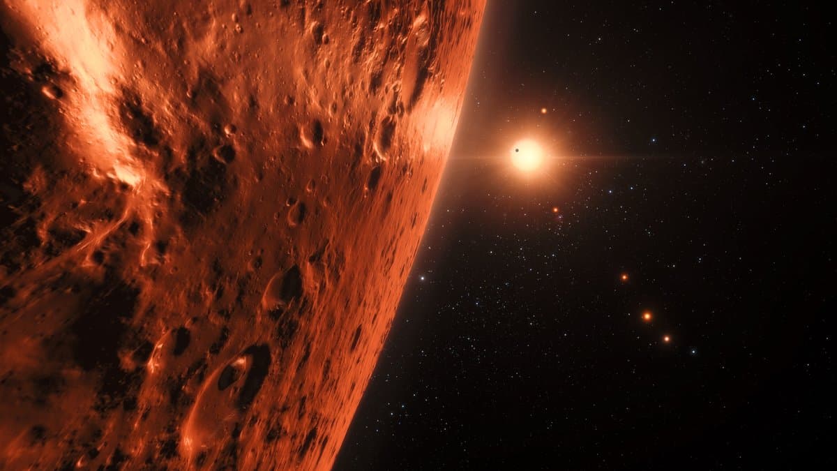 Artist's impression of the TRAPPIST-1 system: a cratered rocky planet lit deep orange in the foreground, with the small red dwarf star and several sibling planets strung out in the distance.
