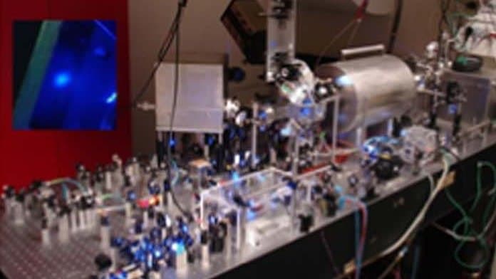 An optical bench crowded with mirrors and lenses lit by blue laser light, running up to a cylindrical vacuum chamber; an inset panel at upper left shows a small cloud of laser-cooled strontium atoms glowing blue.