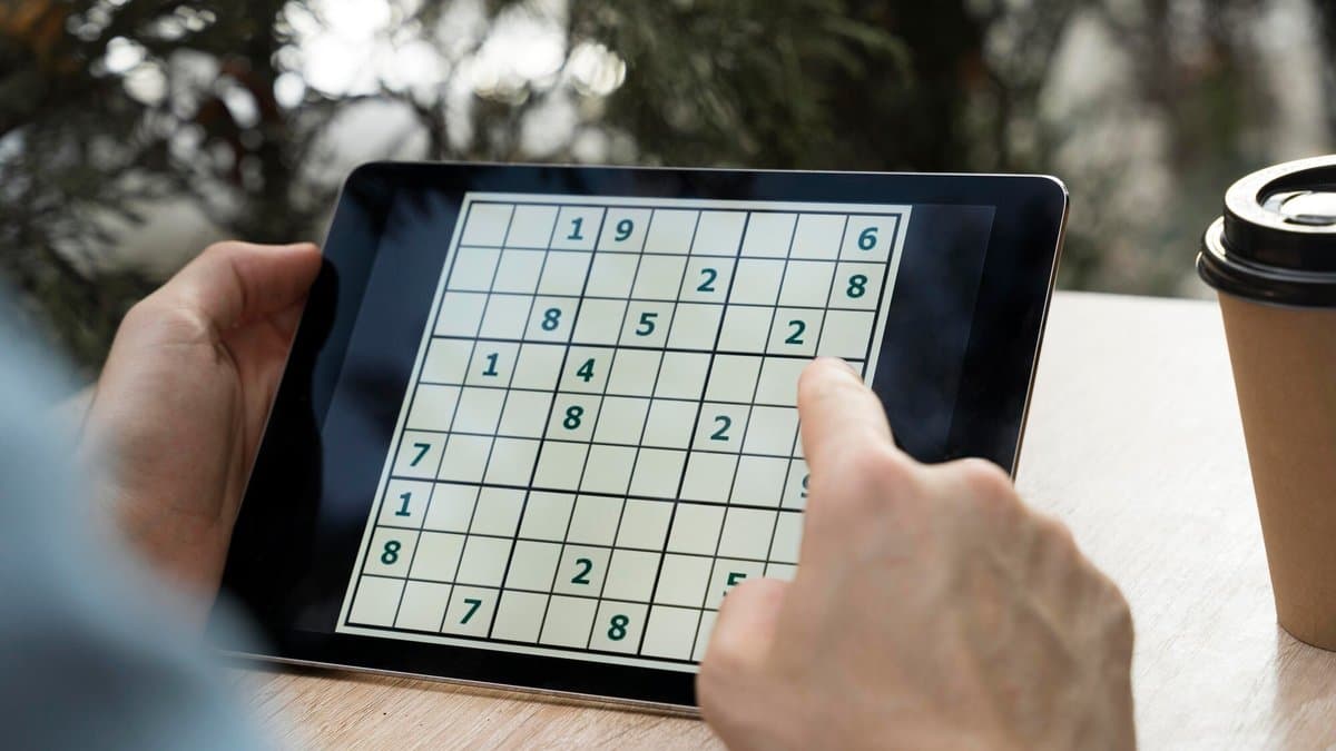A person's hands hold a tablet computer showing a partly filled sudoku grid, resting on a wooden table next to a takeaway coffee cup.