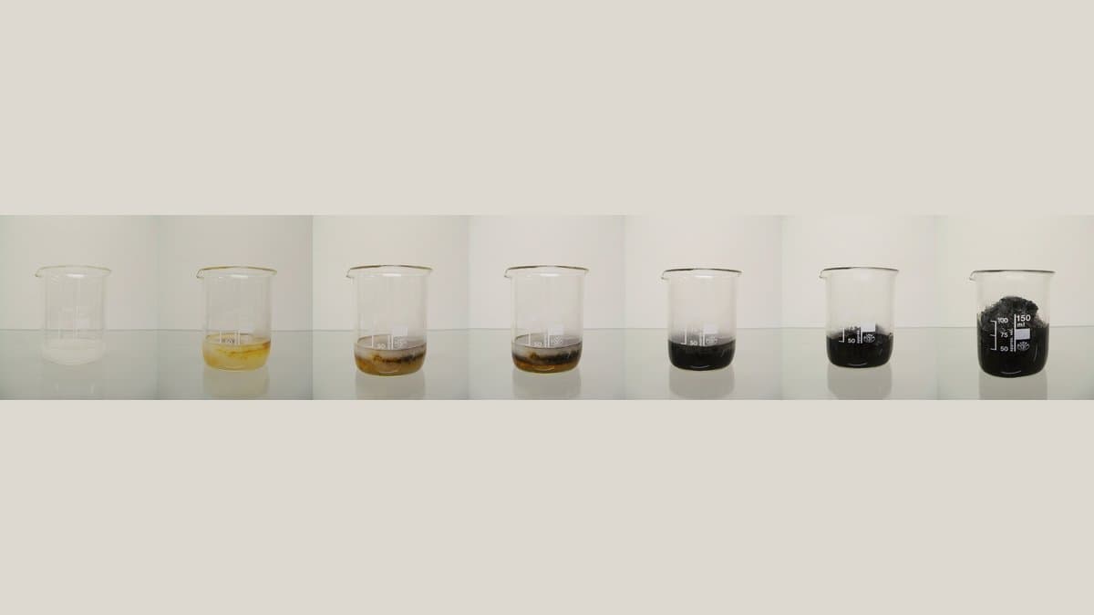 Seven glass beakers photographed in sequence: clear liquid on the left darkens through yellow and brown to a solid black column of carbon on the right.