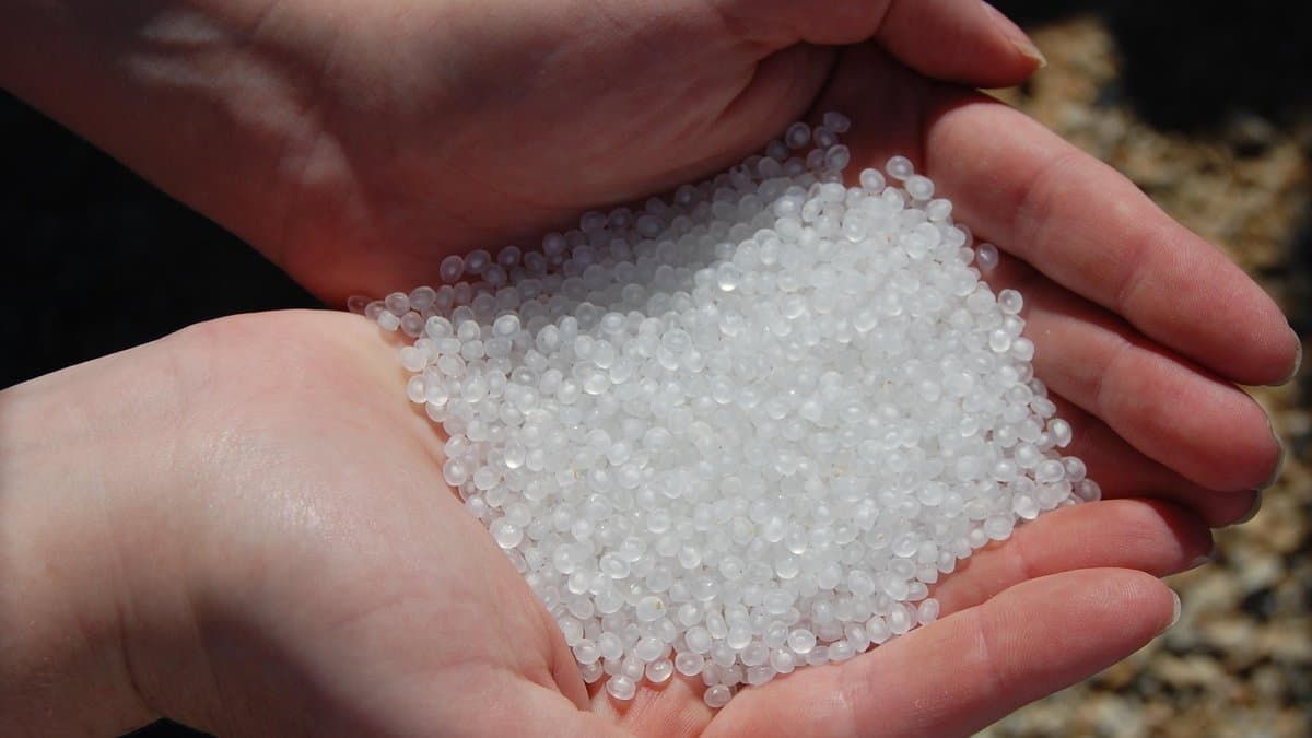Two cupped hands holding a heap of small translucent white plastic pellets, with gravel in the background.